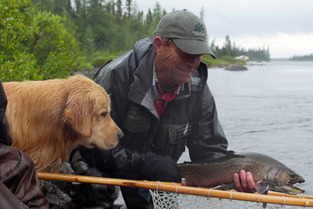 Man holding trout above landing net with dog looking. 
